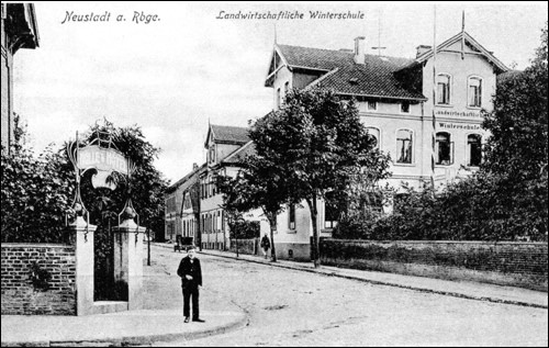 Schwarz-weiß-Postkarte um 1900: Blick auf die ›Landwirtschaftliche Winterschule‹ in Neustadt am Rübenberge; mehrstöckiges Schulgebäude an einer baumgesäumten Straße, im Vordergrund ein Mann im Anzug vor einem Torbogen.