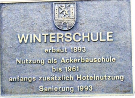 Bronzetafel mit Stadtwappen und erhabener Schrift: ‚Winterschule – erbaut 1893, Nutzung als Ackerbauschule bis 1961, anfangs zusätzlich Hotelnutzung, Sanierung 1993‘.