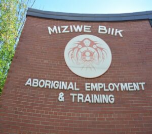 Backsteinwand mit kreisförmigem Logo und Schriftzug ‚Miziwe Biik – Aboriginal Employment & Training‘, Bildungszentrum der First Nations.