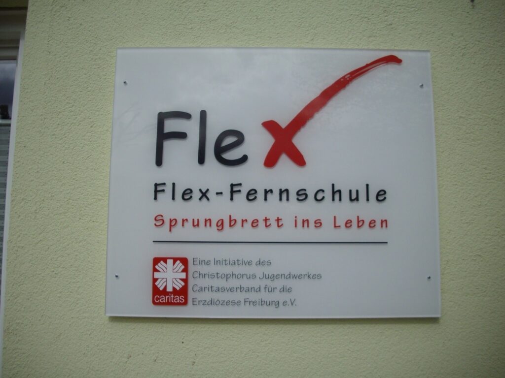 Weißes Wandschild mit Logo der Flex-Fernschule: schwarzer Schriftzug ‚Fle‘, rotes Häkchen als ‚x‘, Slogan ‚Sprungbrett ins Leben‘ sowie Caritas-Logo und Hinweis auf die Trägerschaft durch das Christophorus Jugendwerk und den Caritasverband Freiburg.