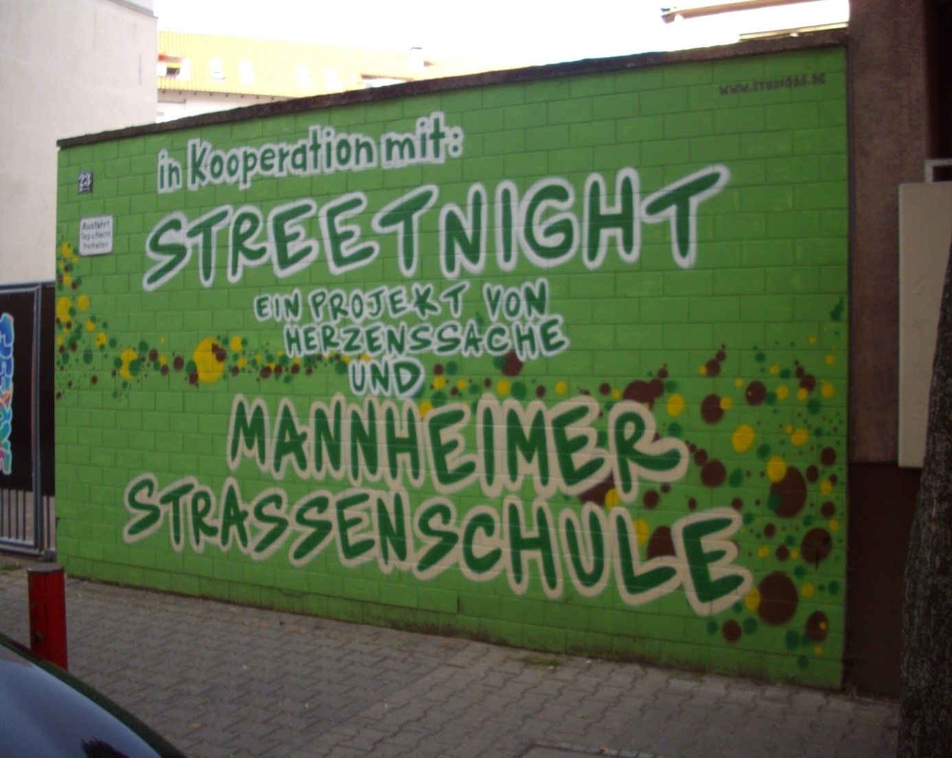 Grün gestrichene Ziegelwand am Straßenrand, gesprayt in weißer Graffiti-Schrift: ‚in Kooperation mit: STREETNIGHT – ein Projekt von Herzenssache und Mannheimer Straßenschule‘; die Wand ist mit gelben, braunen und grünen Farbflecken verziert, davor ein gepflasterter Gehweg.