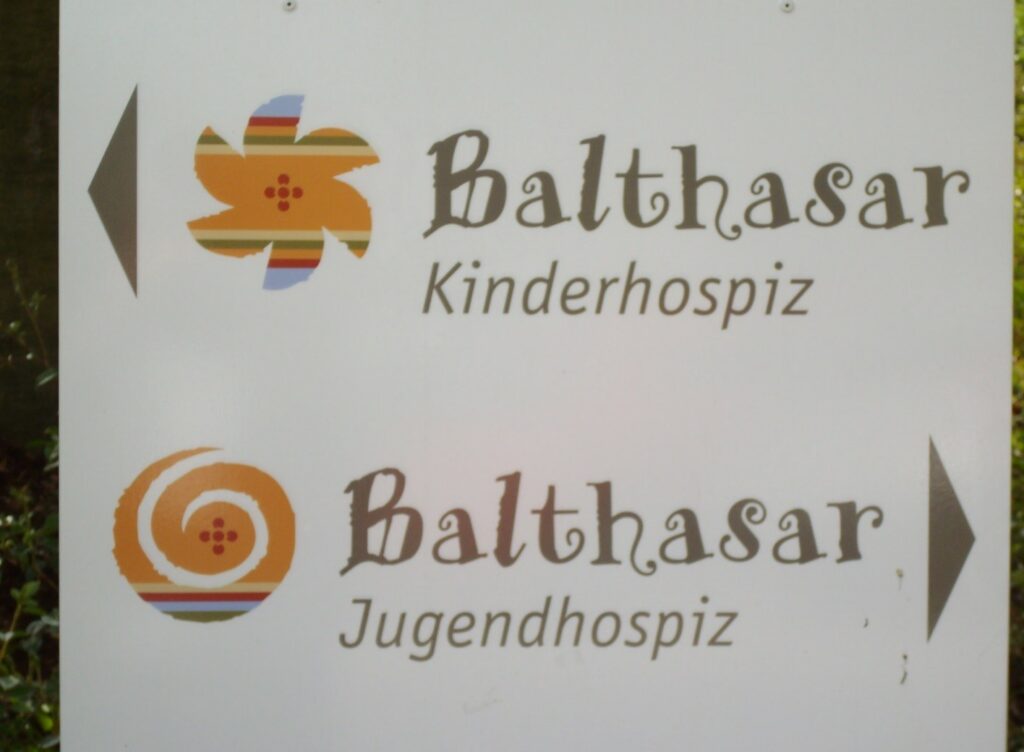 Weißes Hinweisschild mit farbigen Logos und Pfeilen: oben orange-gestylte Blume neben Schriftzug ‚Balthasar Kinderhospiz‘, unten Spirale neben ‚Balthasar Jugendhospiz‘; Pfeile zeigen in entgegengesetzte Richtungen.