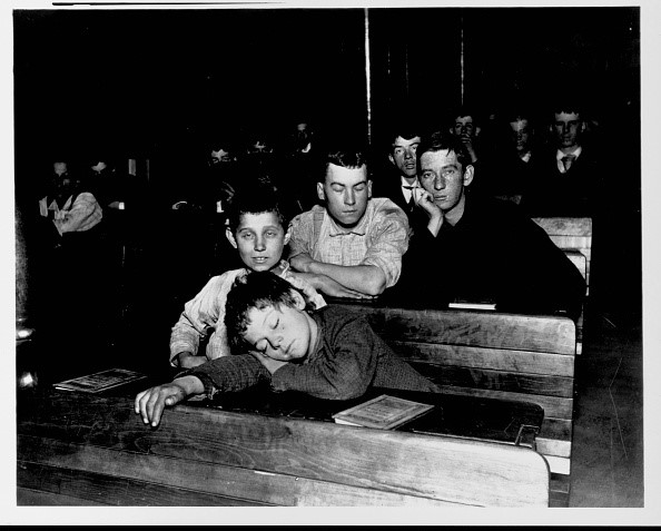 Schwarz-weiß-Foto um 1900: erschöpfte Jungen in einer US-Nachtschule; einer schläft mit dem Kopf auf der hölzernen Bank, zwei weitere stützen müde das Kinn, während der Unterricht weiterläuft.
