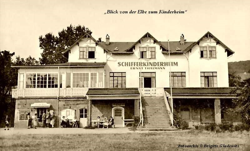 Historische Aufnahme des Schifferkinderheims ›Ernst Thälmann‹ an der Elbe: zweigeschossiges weißes Haus mit Freitreppe, Veranda und spielenden Kindern vor dem Eingang (Blick vom Fluss).