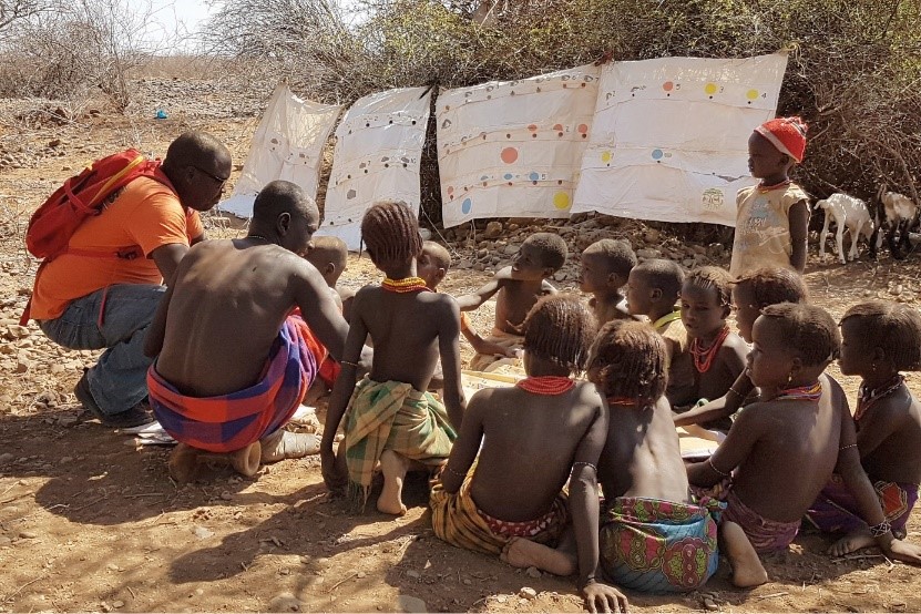 Edwin Changamu besucht Jacob Long’adas mobile Schule in Kenia: er kniet gemeinsam mit einem weiteren Lehrer vor einem Dutzend barfußer Kinder, die im Halbkreis auf steinigem Boden unter freiem Himmel lernen; hinter ihnen hängen große Lernposter mit bunten Punkten.