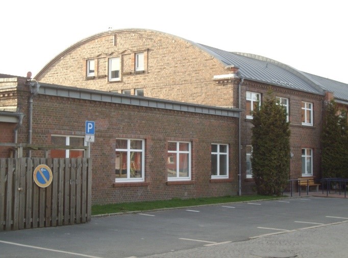 Backstein-Komplex der Produktionsschule mit tonnenförmigem Hallendach, seitlichem Flachbau und Parkplätzen im Vordergrund.