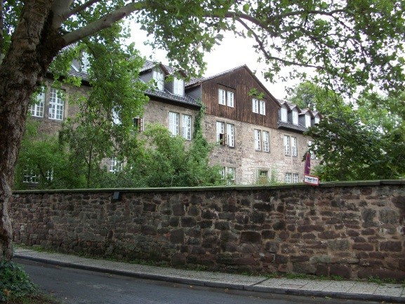 Straßenansicht des langgestreckten Sandstein-Gebäudes der ehemaligen Deutschen Kolonialschule in Witzenhausen; hohe Bruchsteinmauer, dahinter dreigeschossiger Bau mit Satteldach und weißen Sprossenfenstern, von Bäumen umgeben.